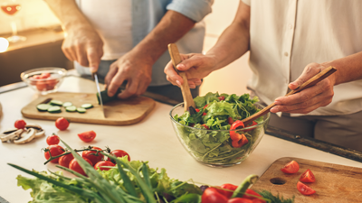 Two people prepare a healthy salad made with lettuce, bell pepper, cucumber, and tomato.