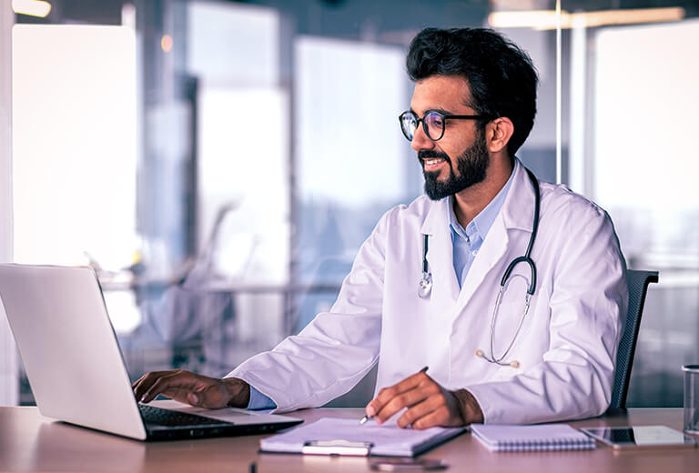 doctor with glasses typing on laptop and writing in notebook